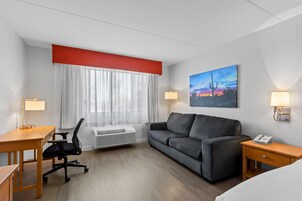 A hotel room with a bed, a desk, a chair, a sofa, an air conditioner, a window with curtains, a painting, and two bedside lamps.
