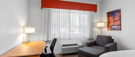 A hotel room with a desk, a chair, a sofa, a bed, an air conditioner, a window with sheer curtains, and two wall-mounted lamps.