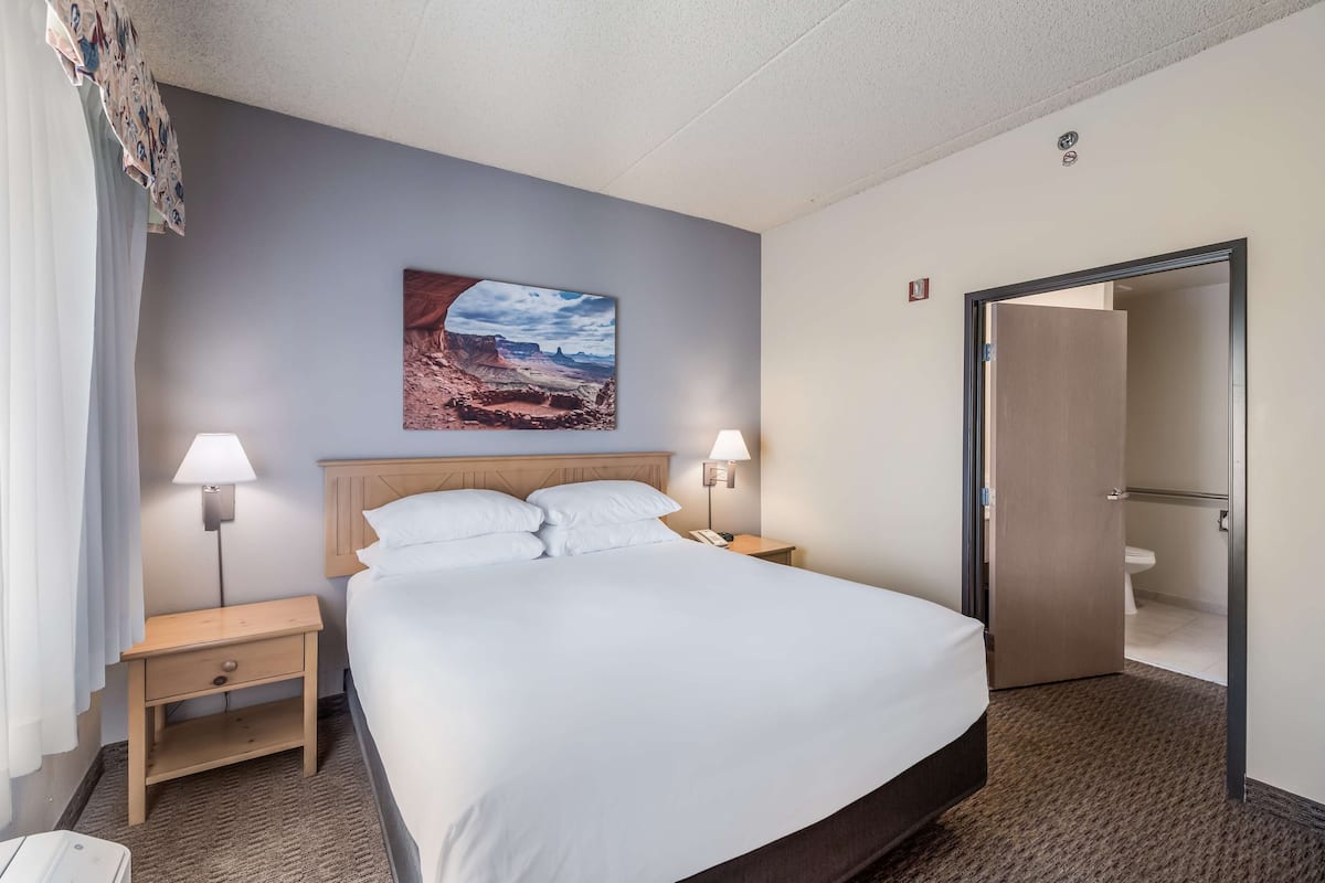 Ein Hotelzimmer mit einem Bett, zwei Nachttischlampen, einem Nachttisch, einem Holz-Kopfteil, einem Bild an der Wand und einer Tür zu einem Badezimmer.