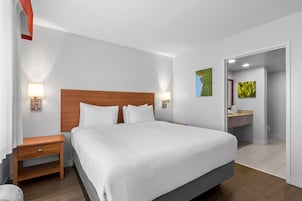 Habitación de hotel con una cama grande, lámparas de noche, una mesita de noche de madera, un baño con lavabo y espejo, y una puerta que da a otra habitación.