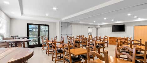 A spacious dining area with multiple wooden tables and chairs, a large window with blinds, and a flat-screen TV mounted on the wall.