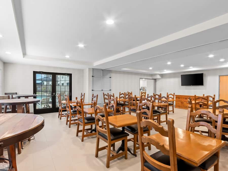 A spacious dining area with multiple wooden tables and chairs, a large window with blinds, and a flat-screen TV mounted on the wall.
