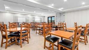 A spacious room with multiple wooden tables and chairs, white walls, and large windows with blinds.