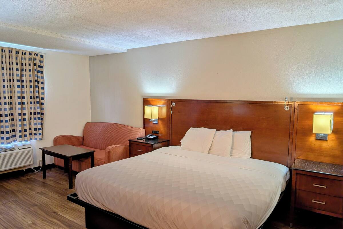 Une chambre d’hôtel avec un grand lit, une tête de lit en bois, une table de chevet avec une lampe, une petite table, une chaise et une fenêtre avec des rideaux.