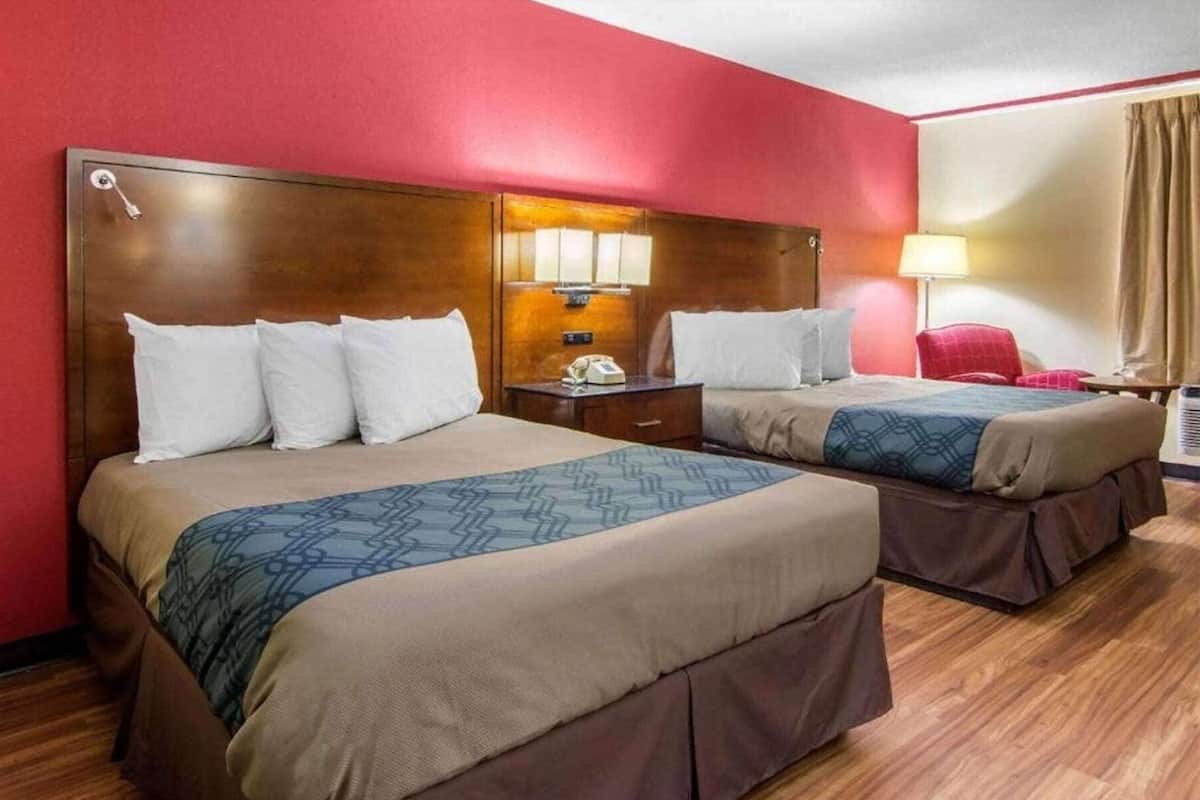 Une chambre d’hôtel avec deux lits, des têtes de lit en bois, un mur rouge, une moquette beige et une fenêtre avec des rideaux beiges.