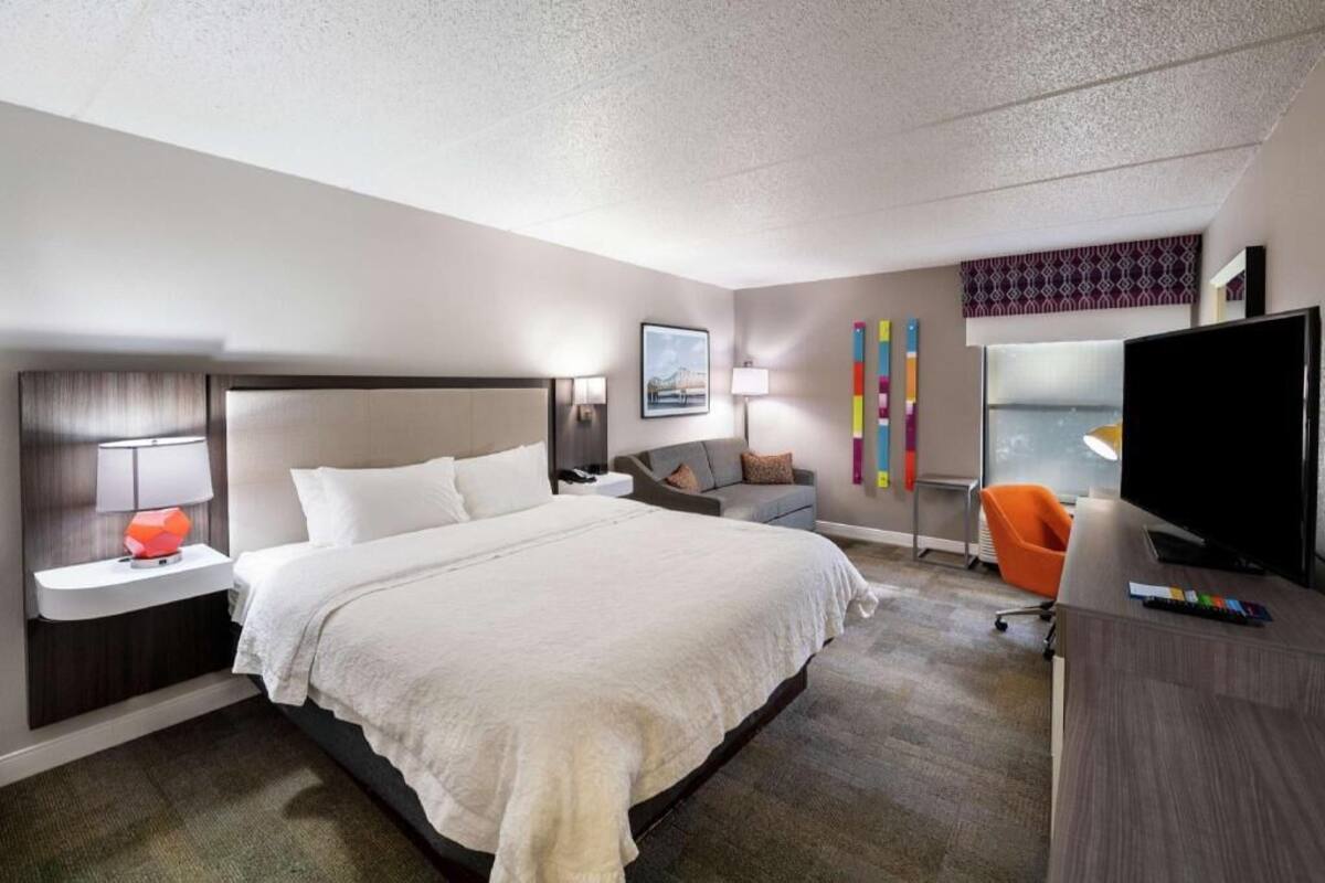 Habitación de hotel con una cama grande, un escritorio con televisor, una silla y una ventana con vistas.
