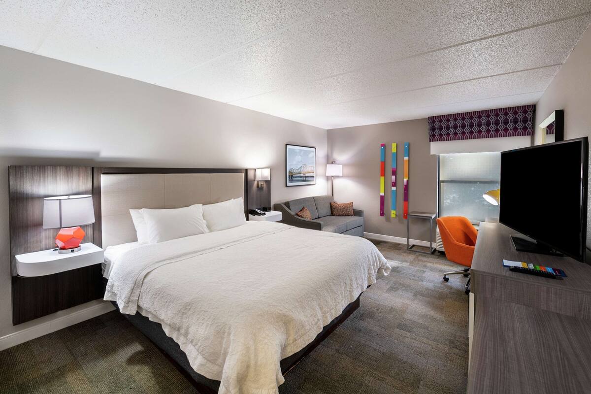 Habitación de hotel con una cama grande, un escritorio con televisor, una silla y una ventana con vistas.
