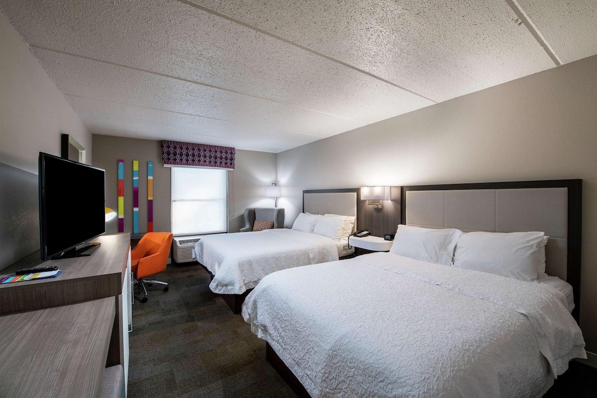 Habitación de hotel con dos camas, un escritorio con televisor, una silla y una ventana con cortinas estampadas.