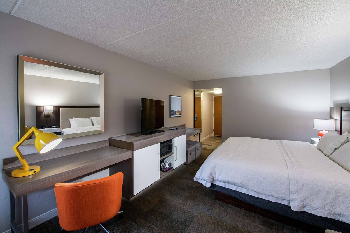 Habitación de hotel con cama, escritorio, silla, televisor y un espejo grande.