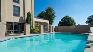 A swimming pool with a clear blue water surface, surrounded by a beige building with a covered patio area and a metal fence. - Hampton Inn Gainesville (Gainesville)