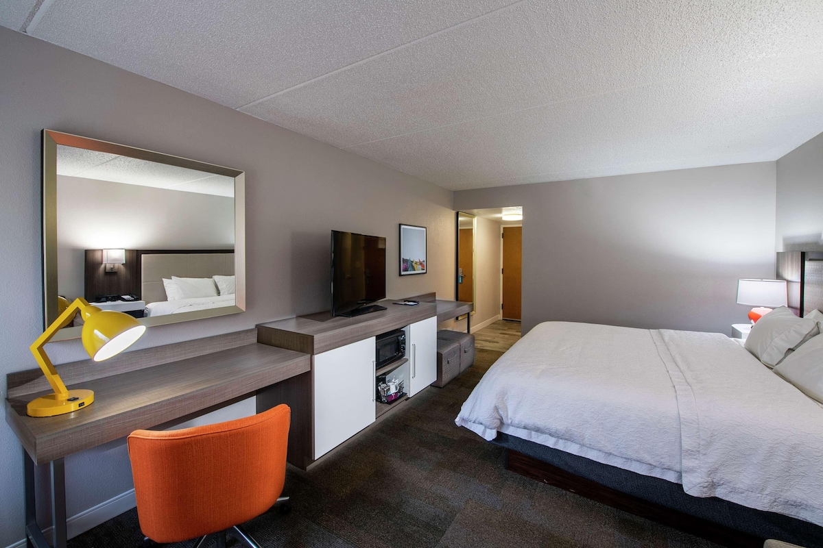 Una habitación de hotel con cama, escritorio, silla, televisión, espejo y microondas.