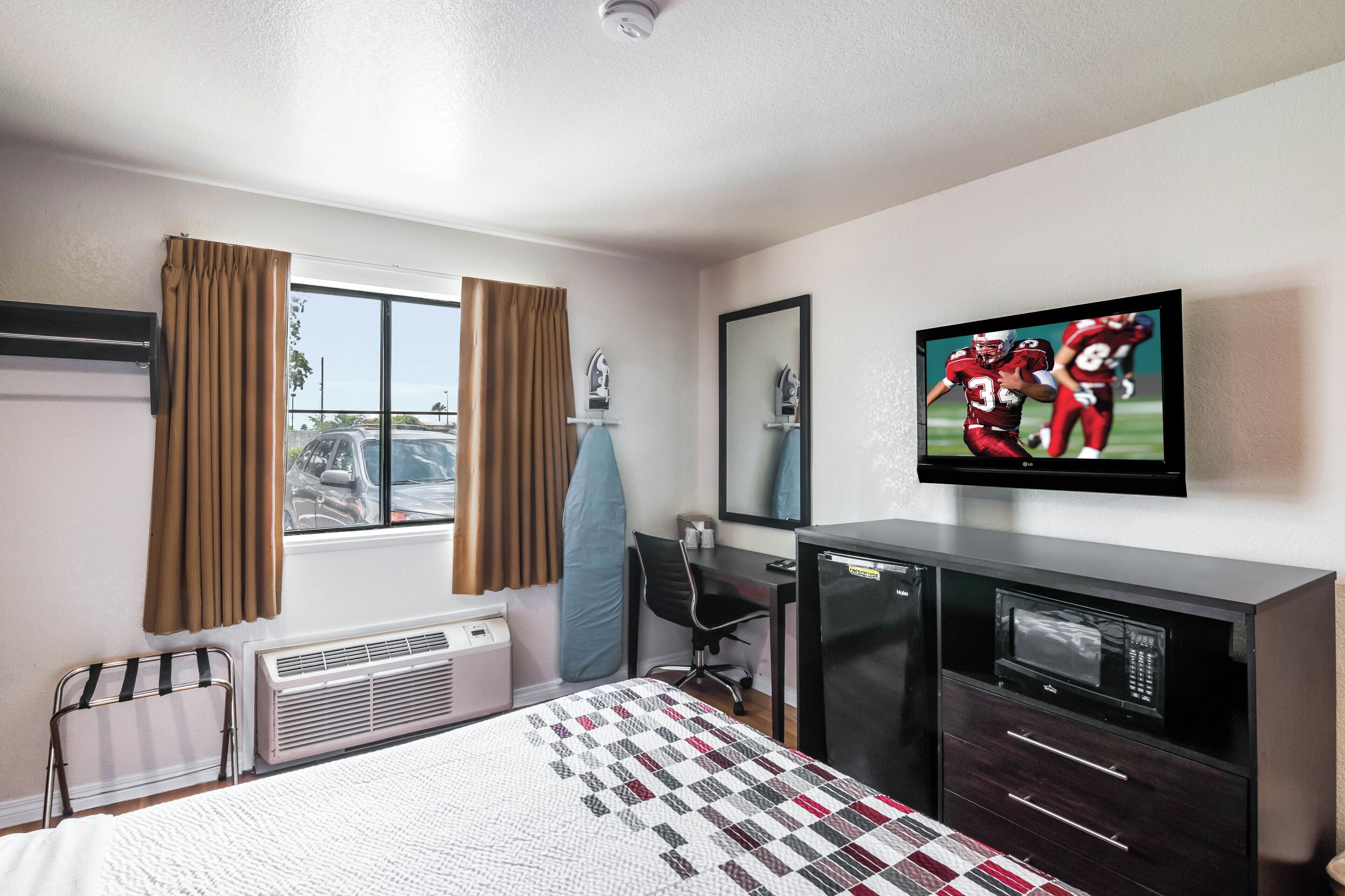 Habitación de hotel con cama, escritorio, televisor, espejo y ventana con cortinas.
