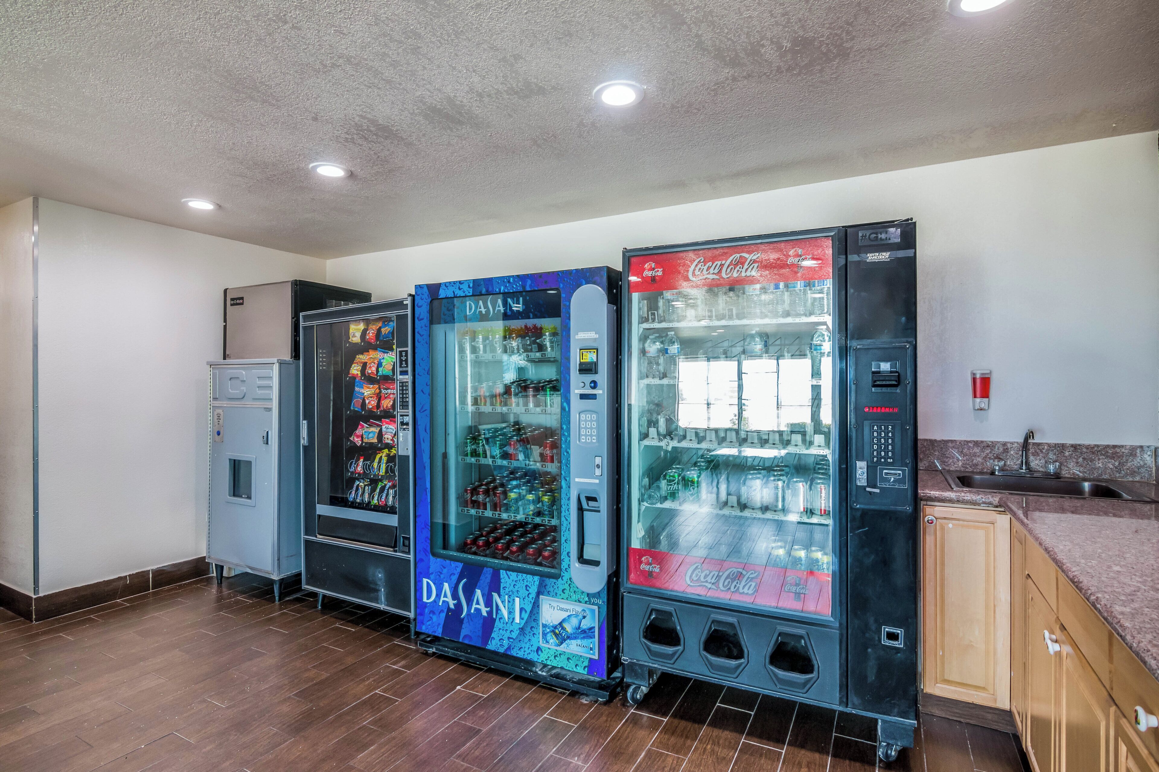 Een rij automaten met diverse drankjes, waaronder Coca-Cola en Dasani water, in een ruimte met houten vloeren en een kitchenette.