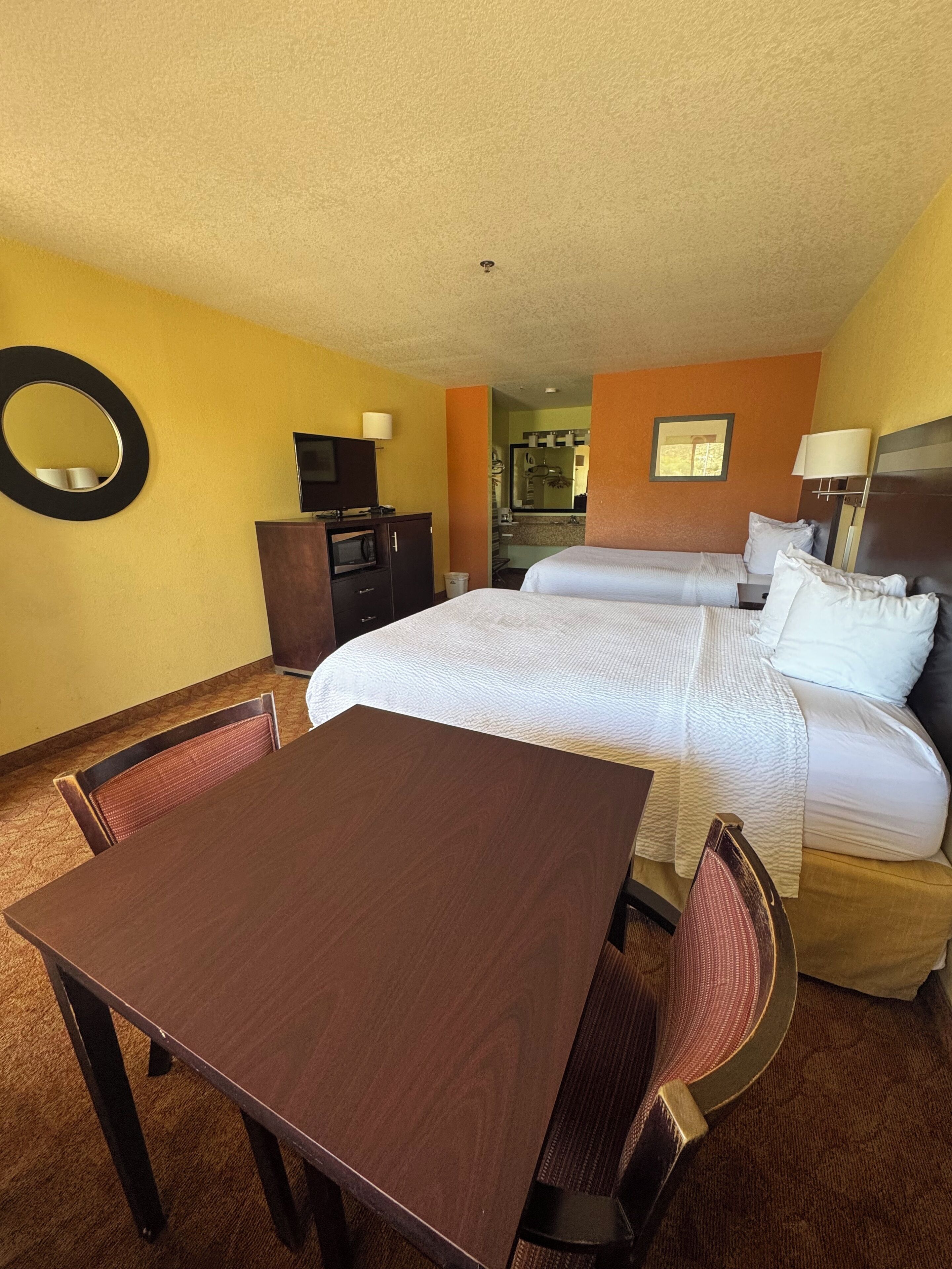 Quarto de hotel com cama, escrivaninha, cadeira, televisão e espelho.