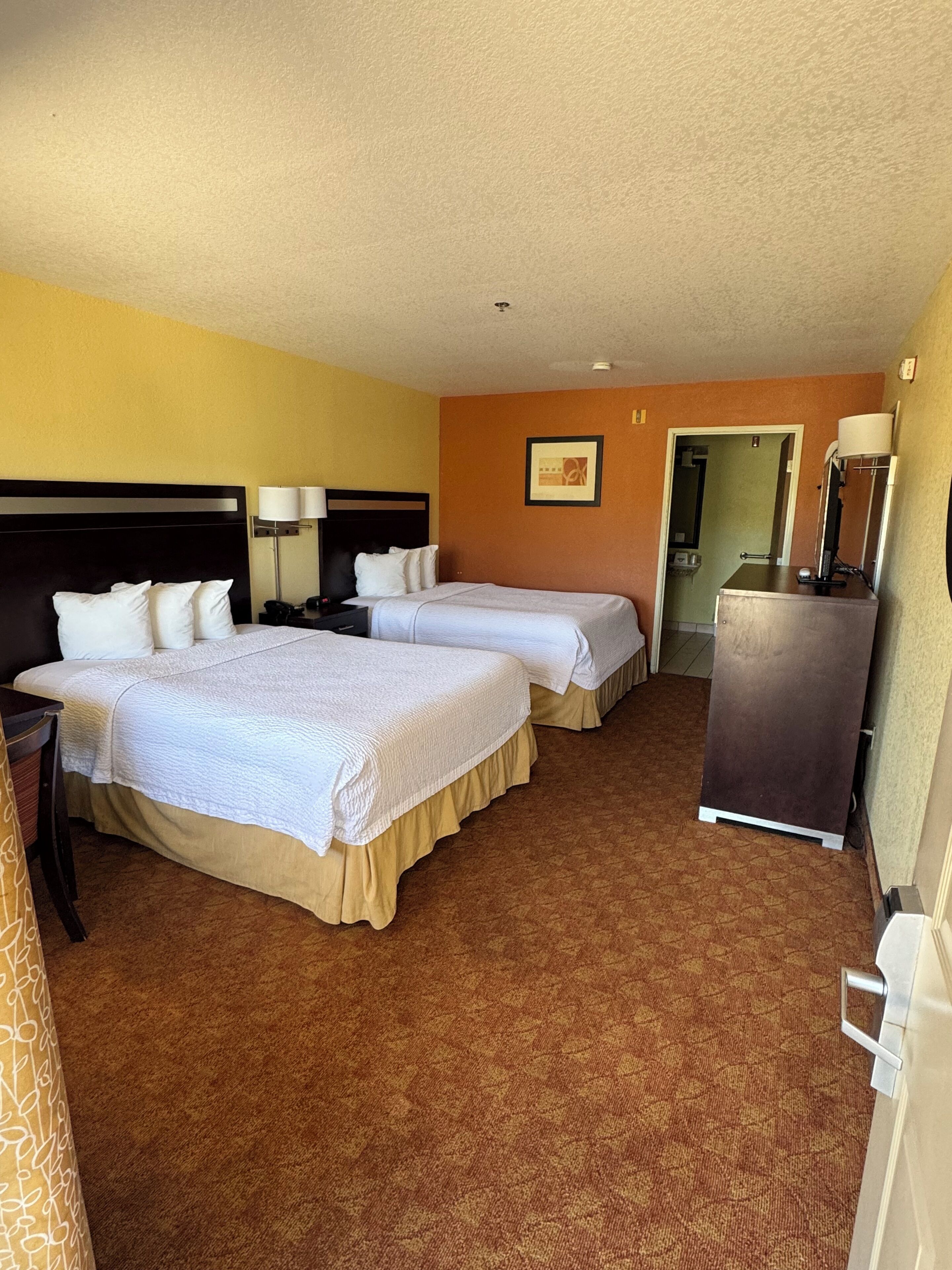 A hotel room with two beds, a desk, a dresser, and a door leading to a bathroom.