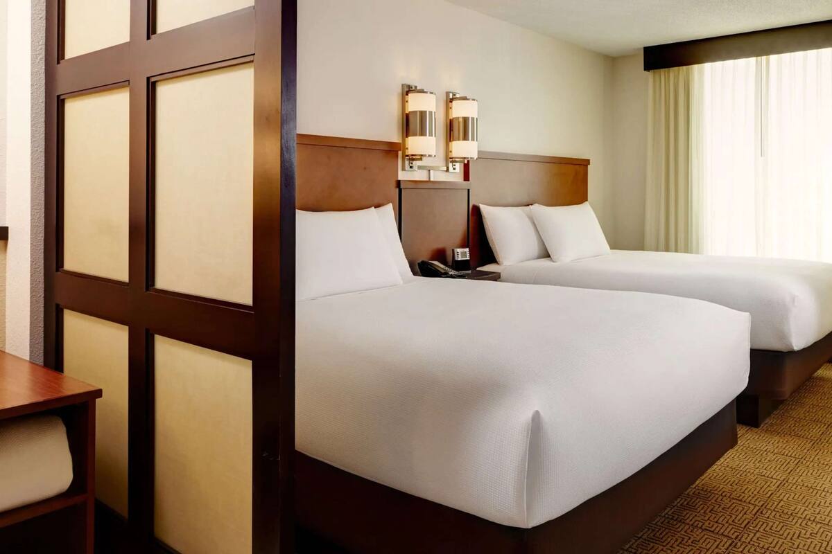 A hotel room with two beds, a wooden headboard, a bedside table with a lamp, and a window with curtains.