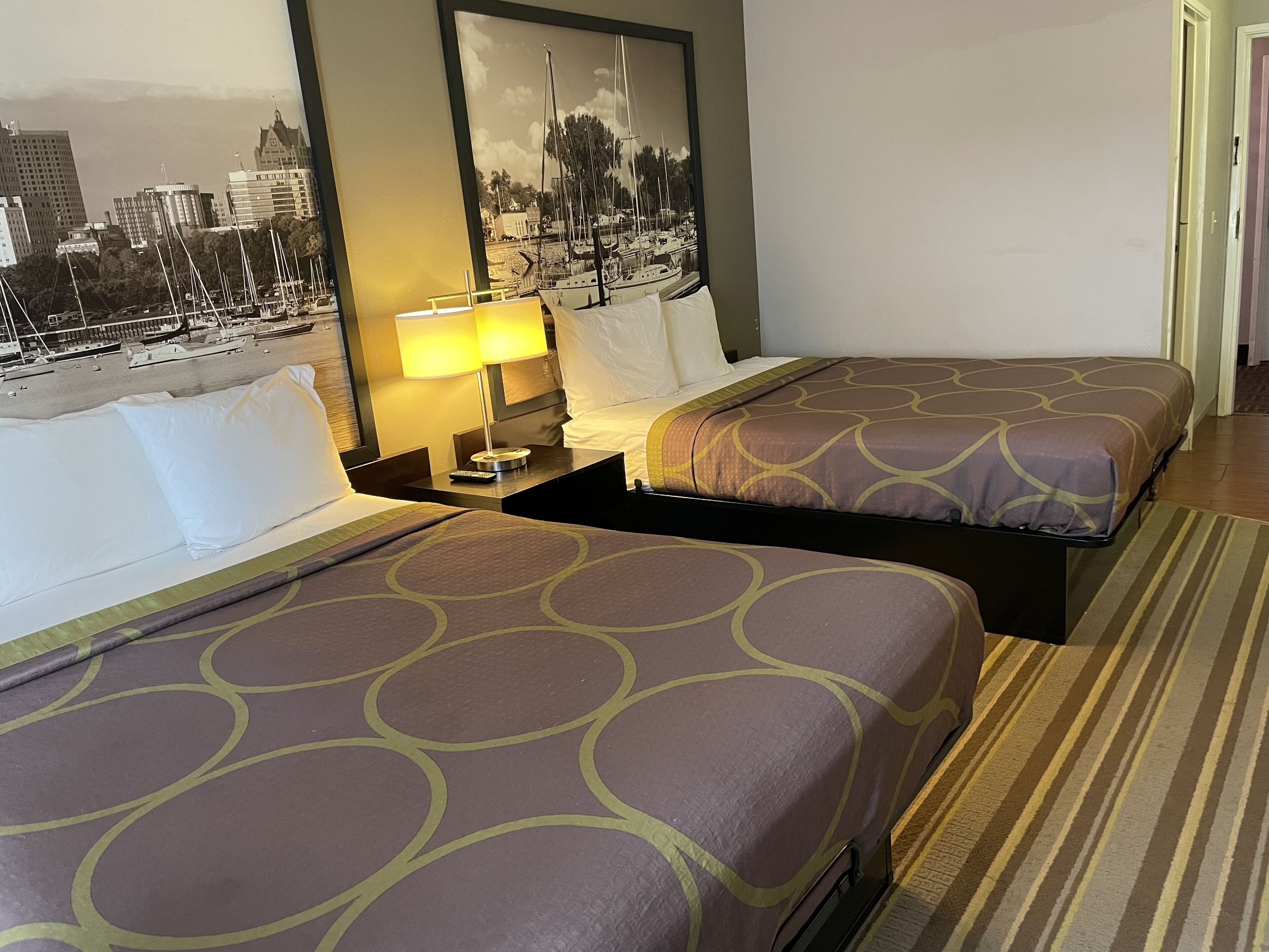 A hotel room with two beds, a nightstand with a lamp, and a cityscape view.