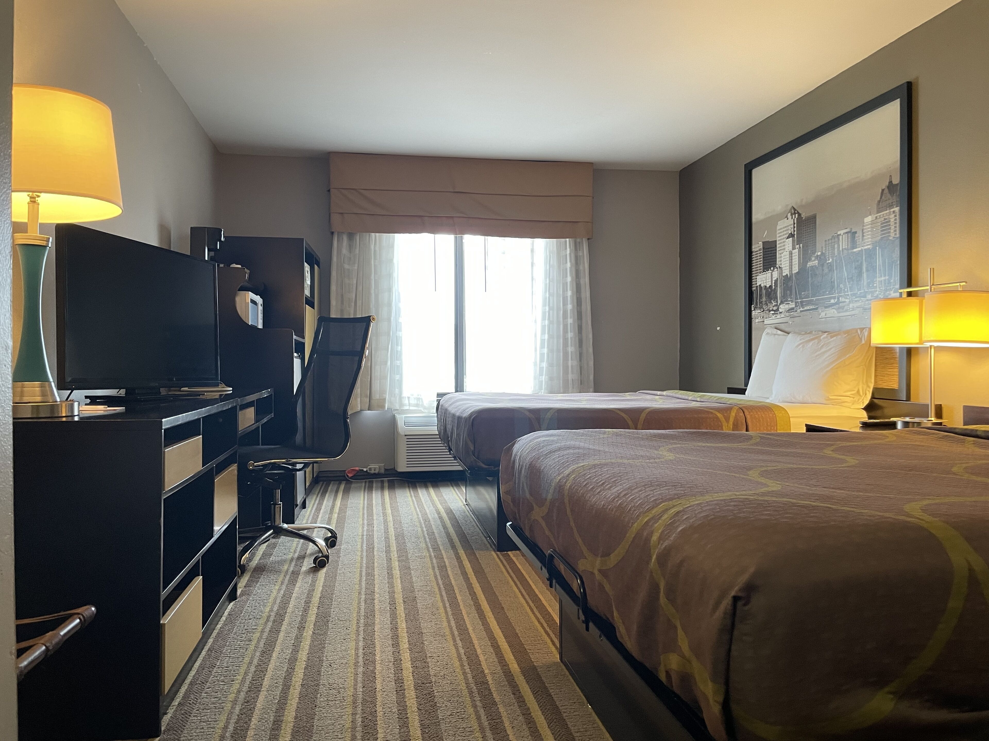 A hotel room with two beds, a desk with a TV, a chair, a lamp, and a large window with curtains.