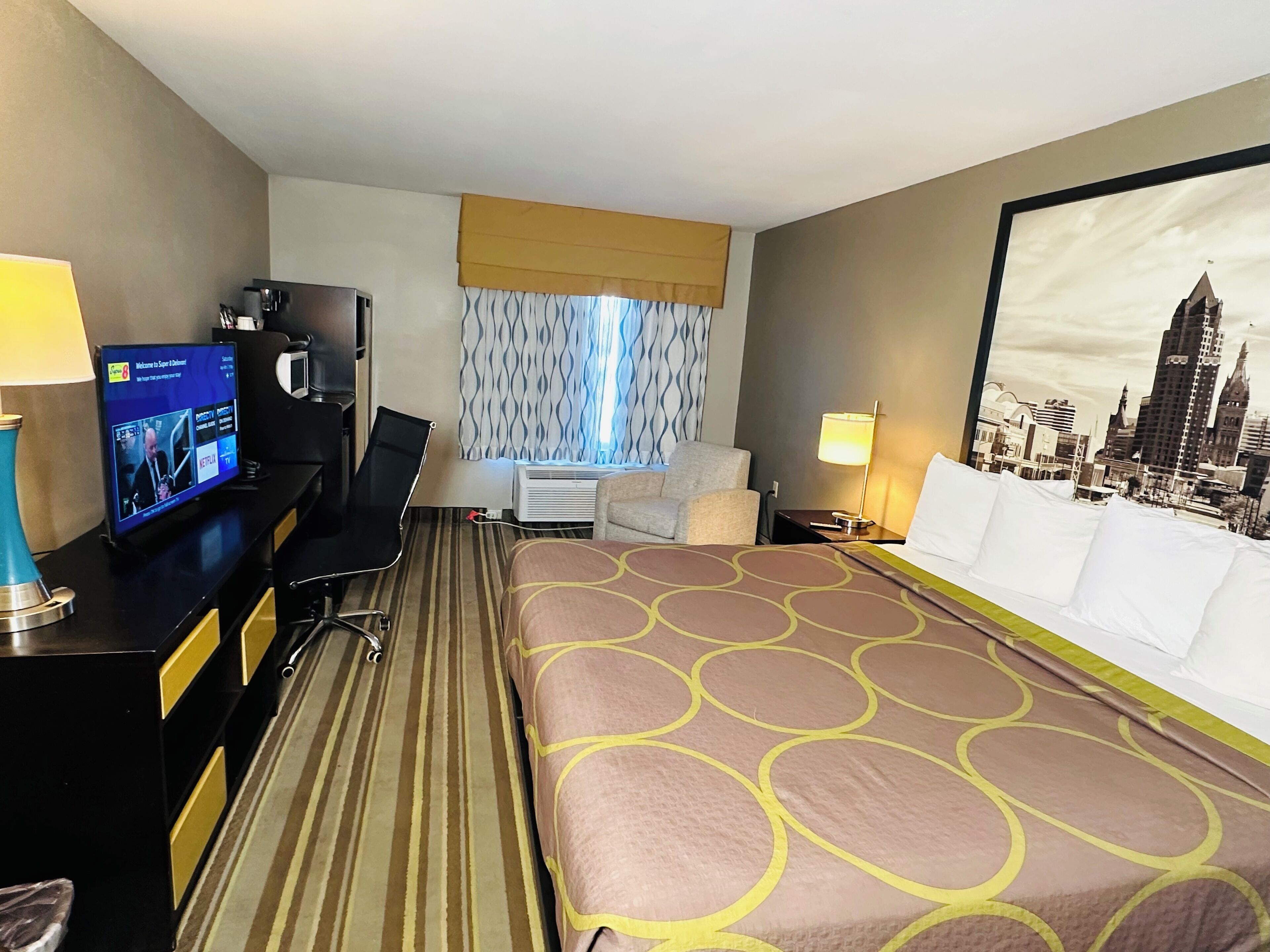 A hotel room with a bed, a desk with a computer, a chair, a window with curtains, and a cityscape view.