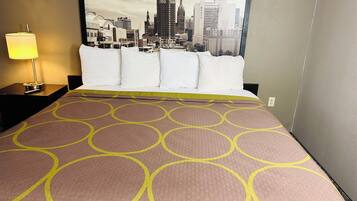 A hotel room with a bed featuring a patterned bedspread, a nightstand with a lamp, and a cityscape view through the window.