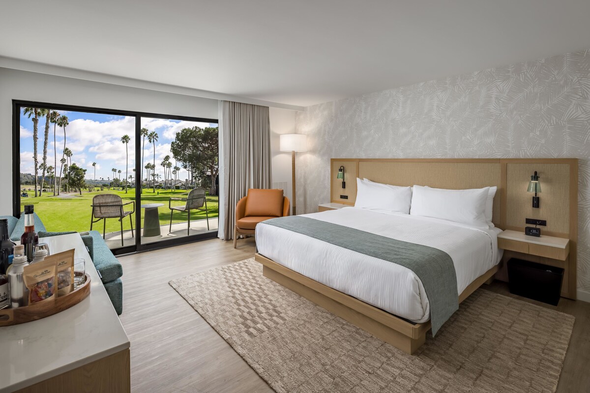 Quarto de hotel com uma cama grande, uma escrivaninha com cafeteira, uma cadeira e vista para um campo de golfe.