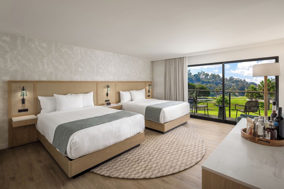Quarto de hotel com duas camas, uma janela ampla com vista para um campo de golfe, um tapete redondo e uma escrivaninha com artigos de higiene pessoal.