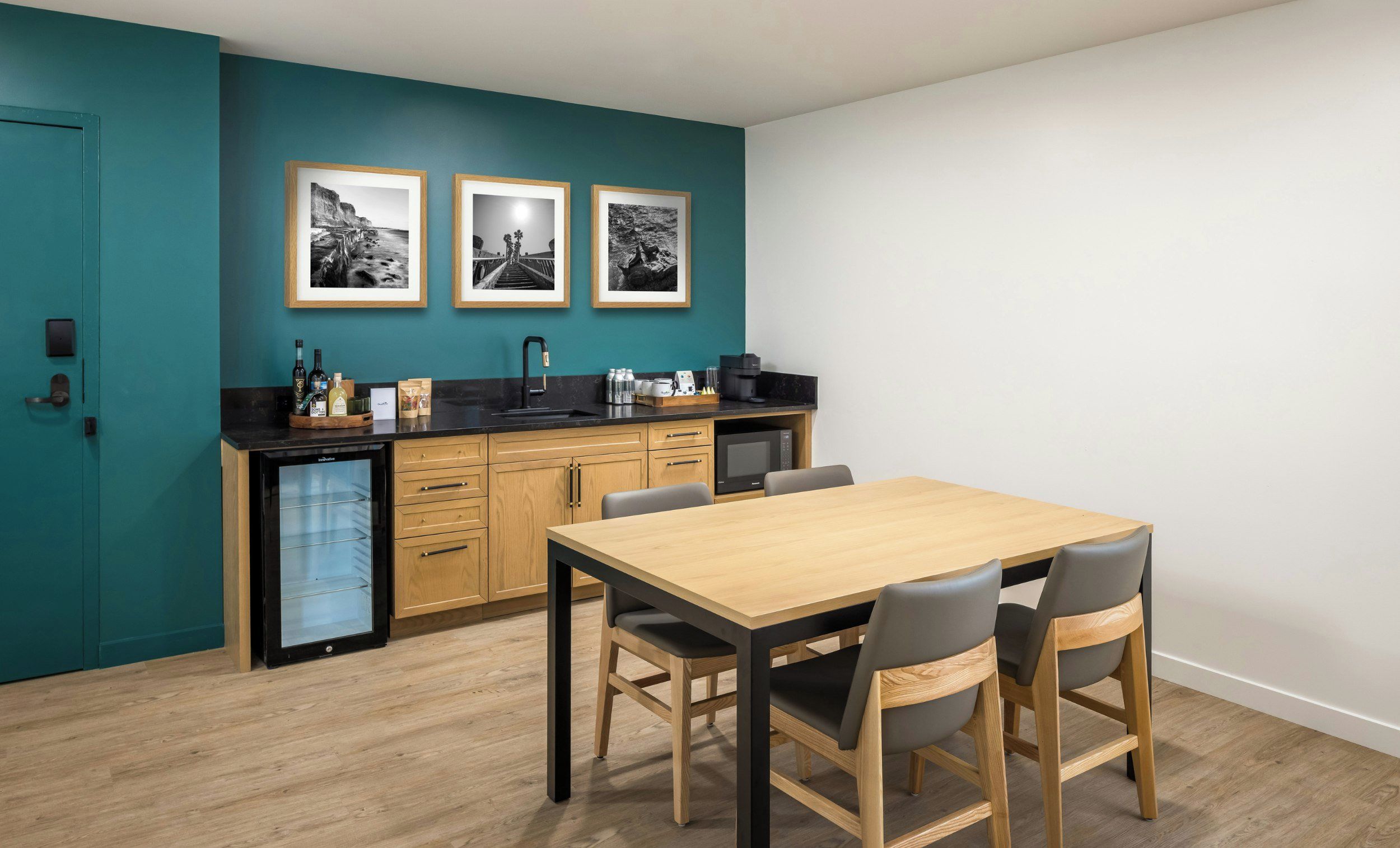 A compact kitchen with a wooden table and chairs, a dishwasher, and a refrigerator. There are three framed pictures on the wall.