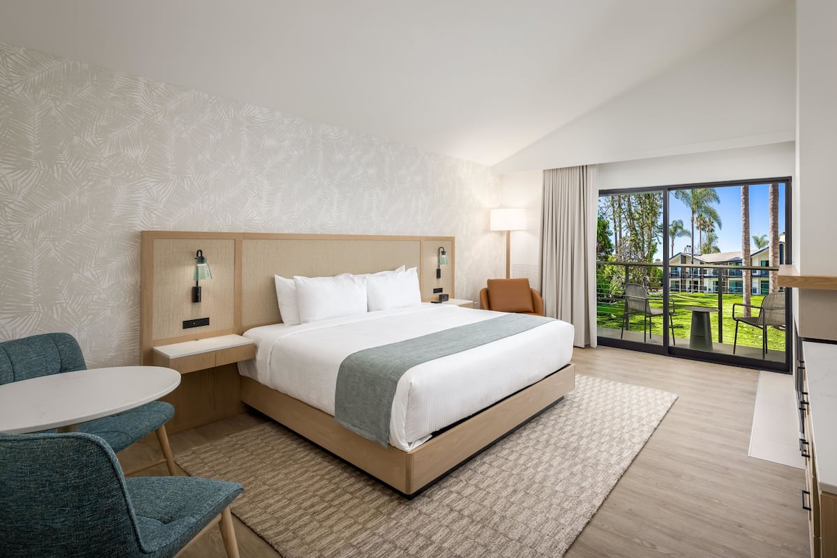 A hotel room with a bed, a desk, a chair, a table, and a view of a garden.