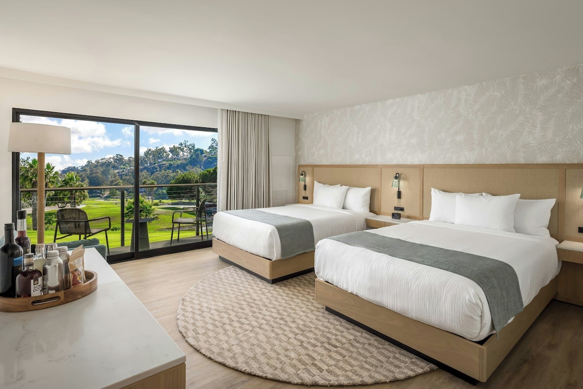 A hotel room with two beds, a large window with a view of a golf course, a round rug, and a small table with bottles and glasses.