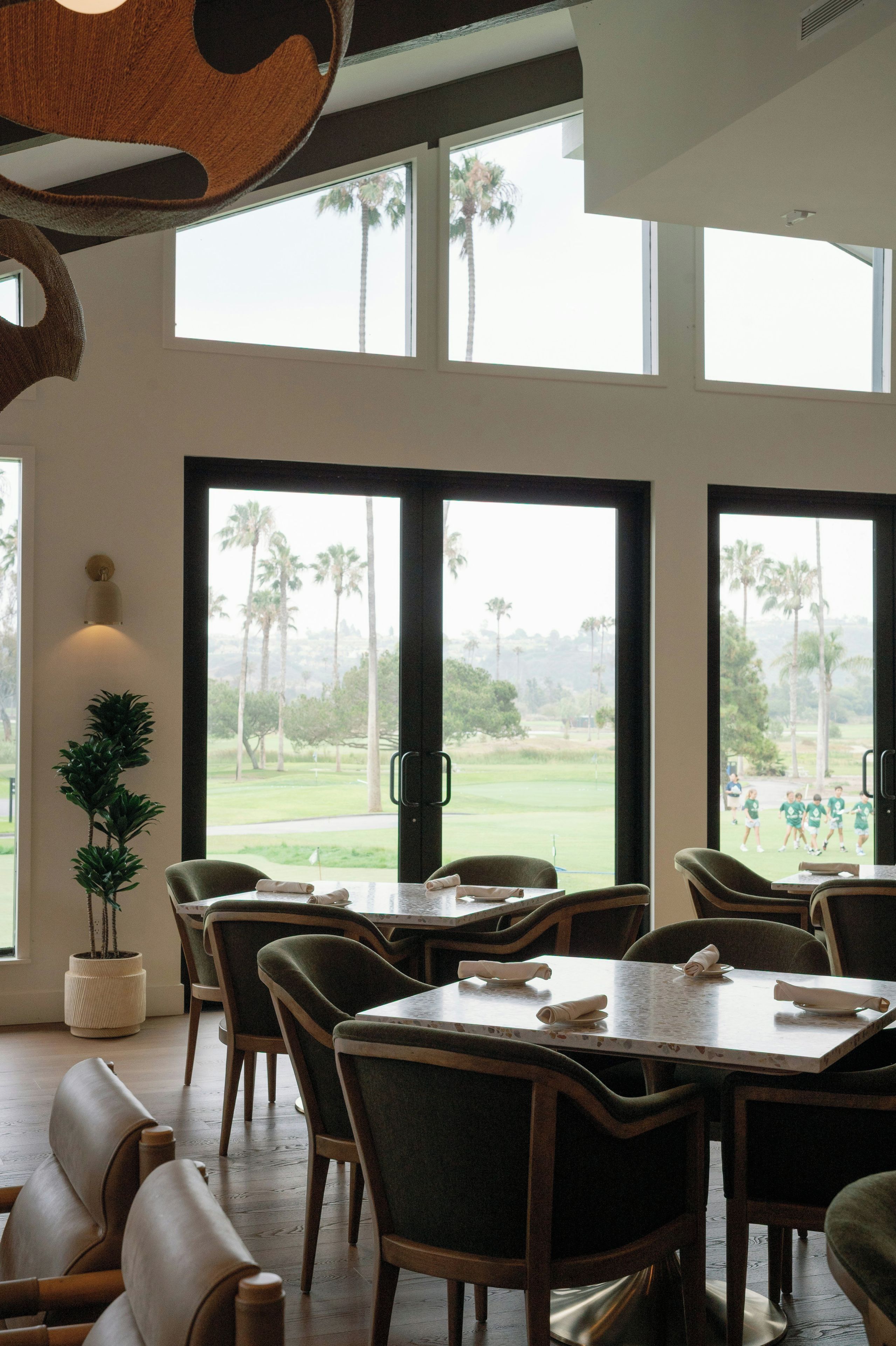 A dining area with tables and chairs, large windows with a view of a golf course, and a potted plant.