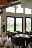 Een eetruimte met tafels en stoelen, grote ramen met uitzicht op een golfbaan, en een kamerplant.