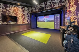 Een golfsimulatieruimte met een groot scherm waarop een golfbaan wordt weergegeven, een gele mat en een golftas.
