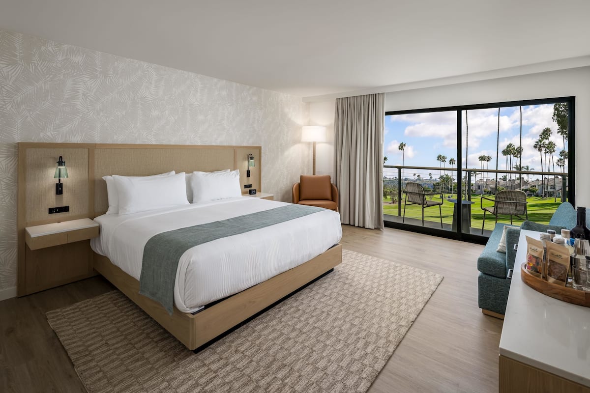 A hotel room with a large bed, a desk with a chair, a view of the outdoors, and a balcony with chairs.