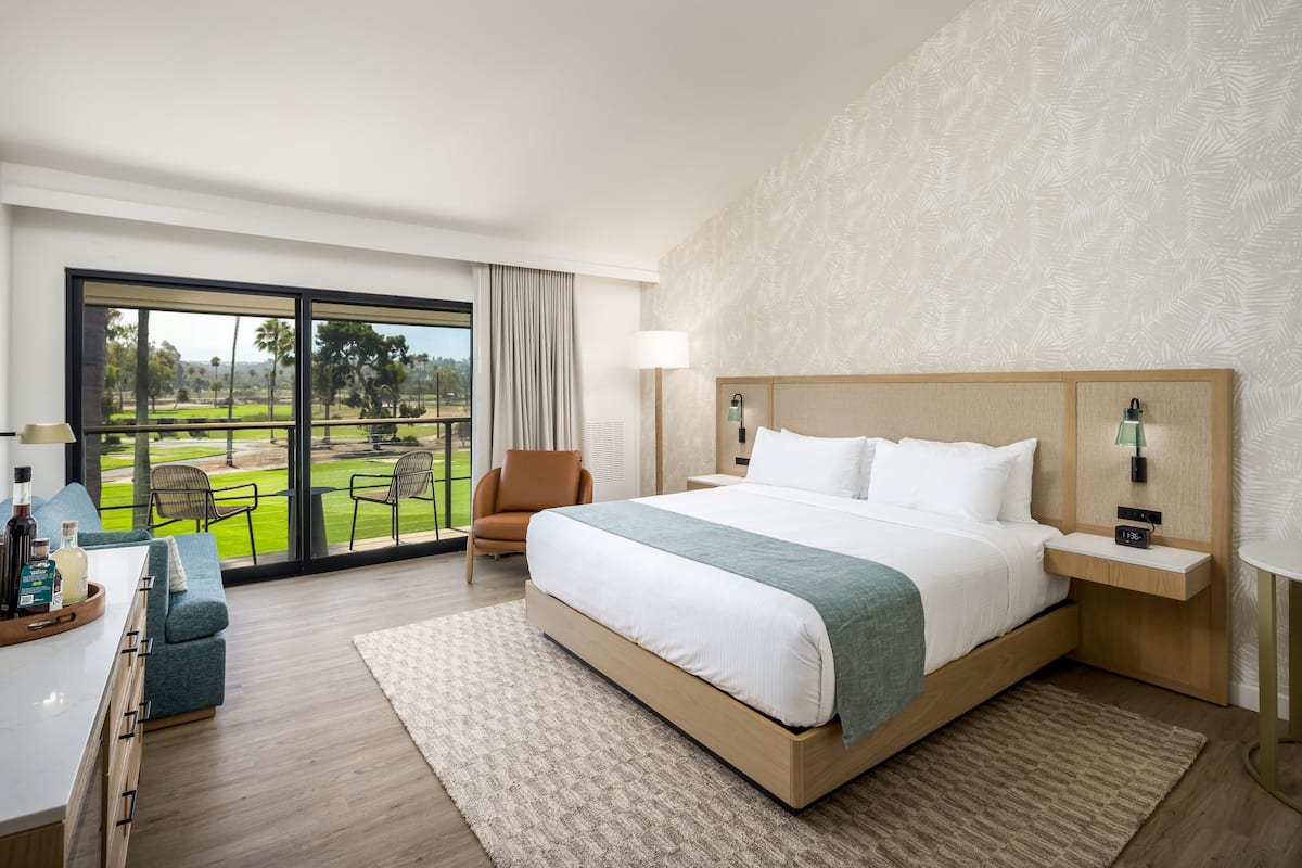 A hotel room with a large bed, a desk with a chair, a window with a view of a golf course, and a nightstand with a lamp.