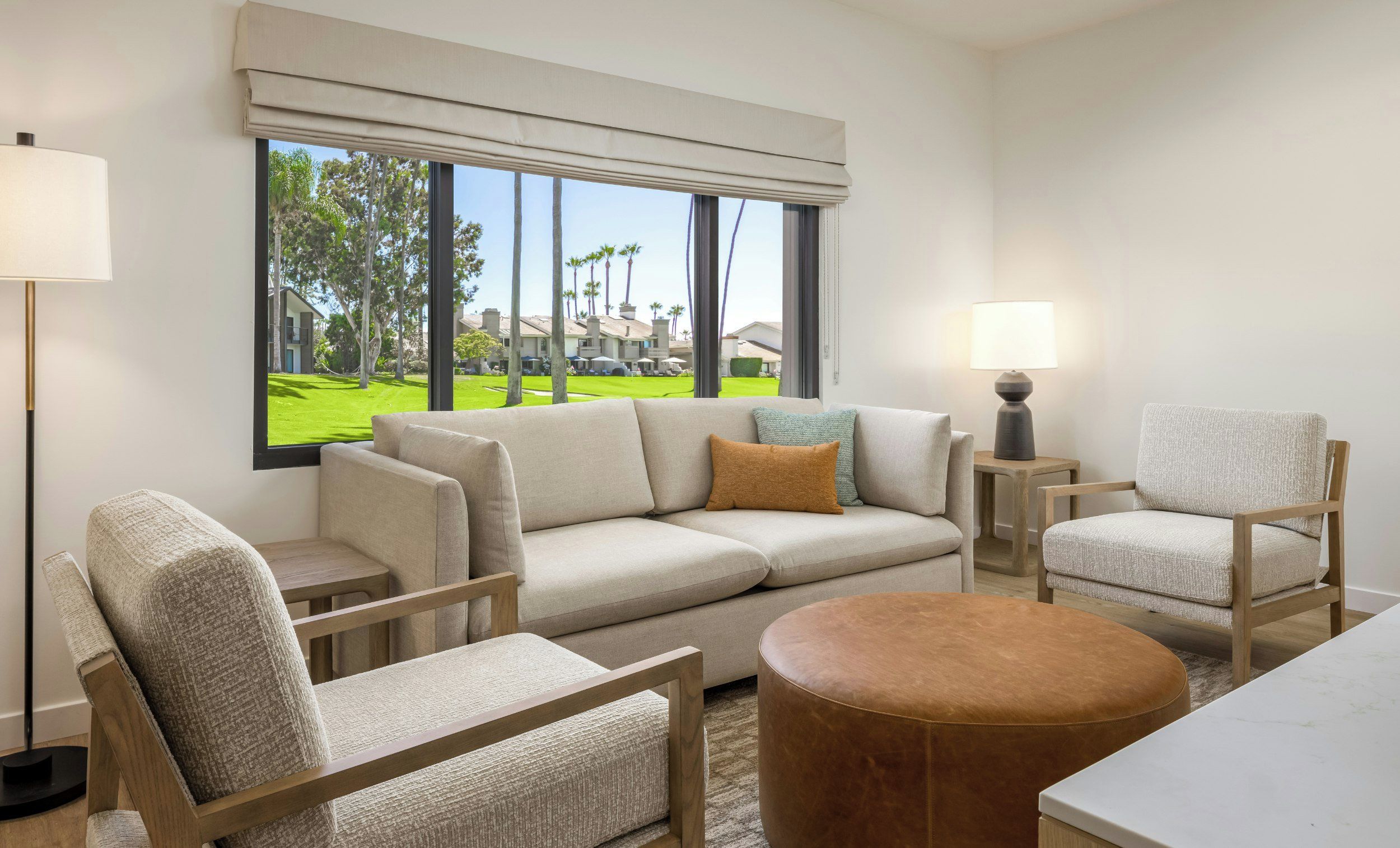 A modern living room with a large window, a sofa, two armchairs, a round ottoman, and a side table with a lamp.