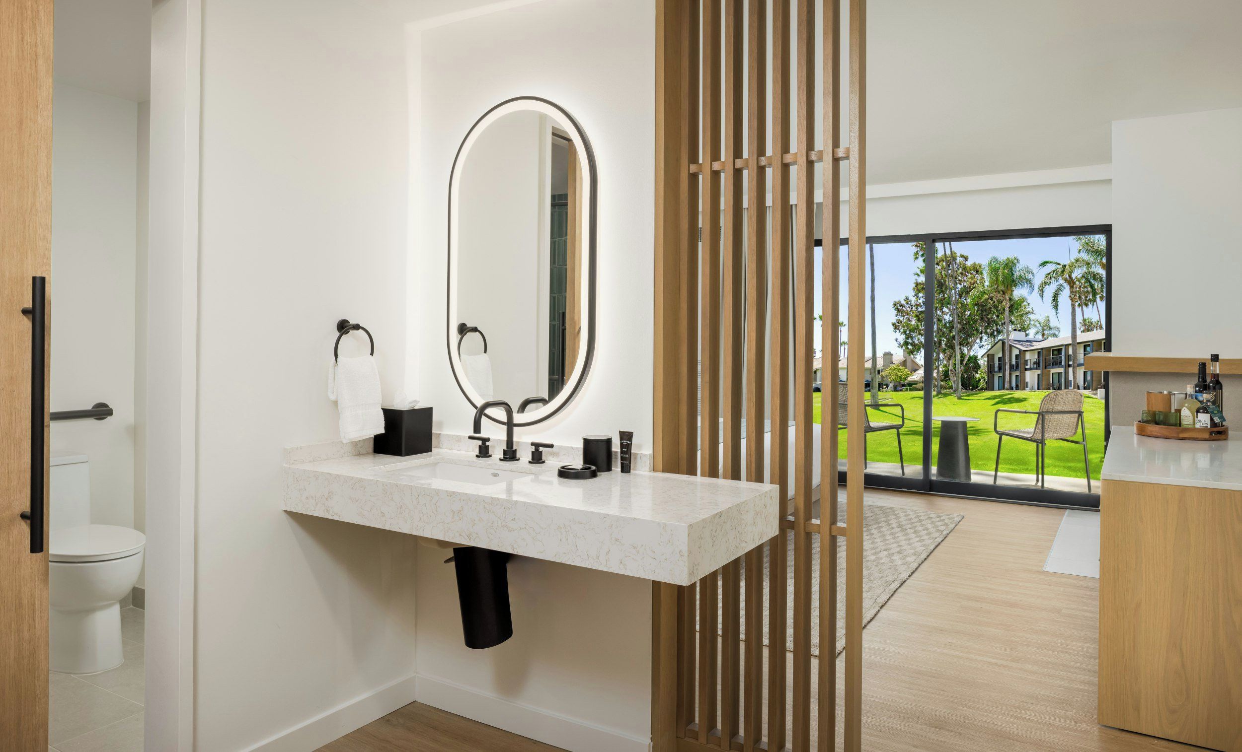 A modern bathroom with a marble sink, a large mirror, and a view of a green outdoor area through a glass door.