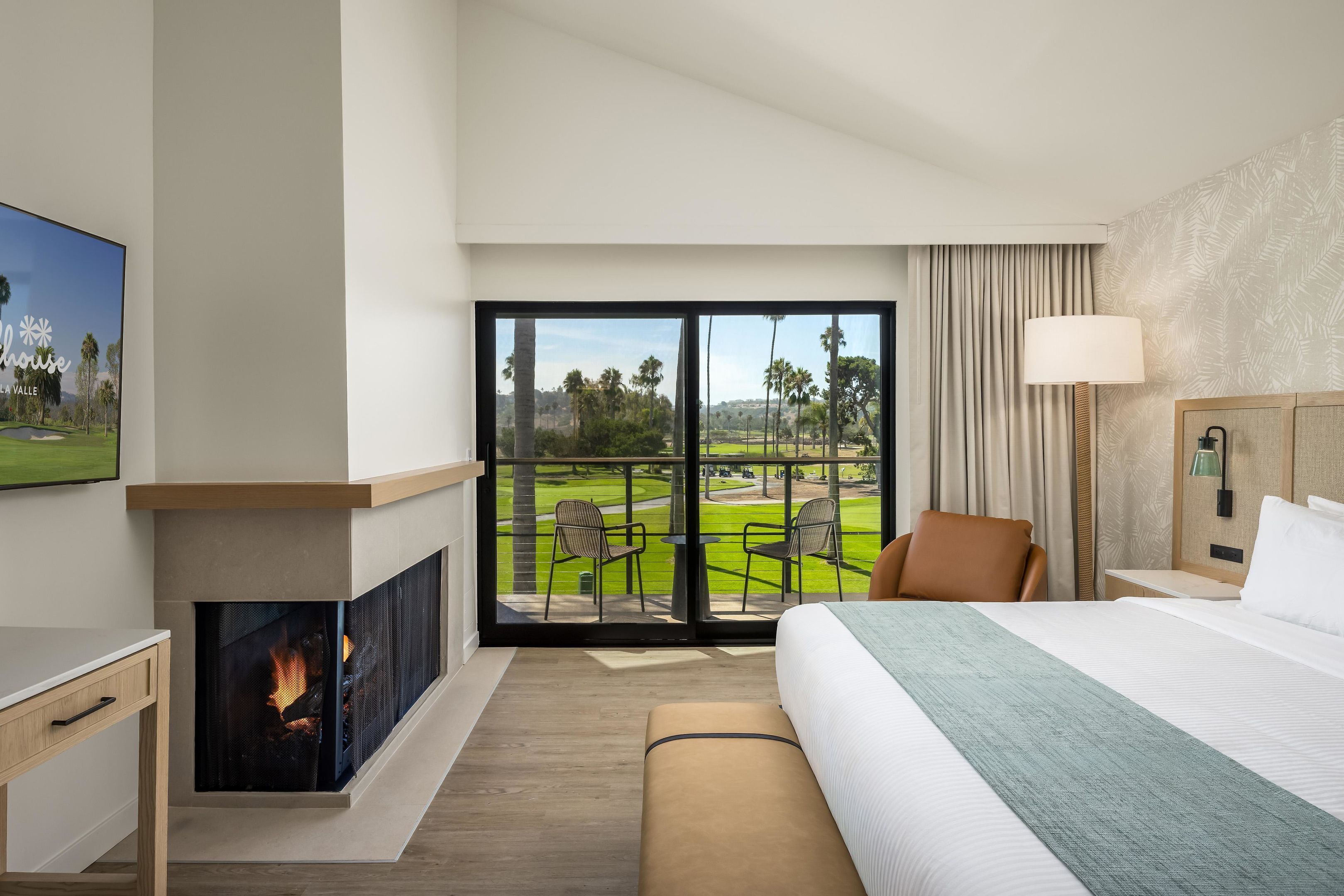 A hotel room with a large bed, a fireplace, a television, and a balcony with a view of a golf course.