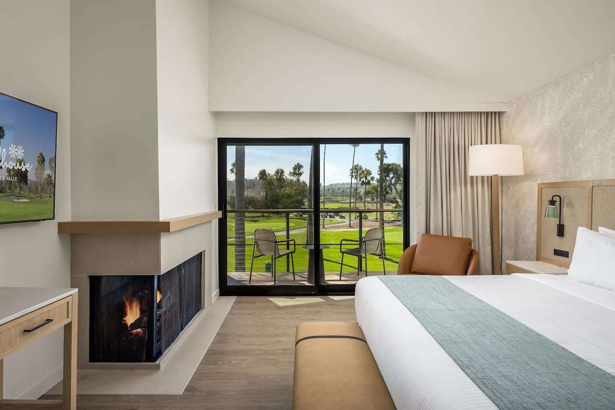 A hotel room with a large bed, a fireplace, a television, and a balcony with a view of a golf course.