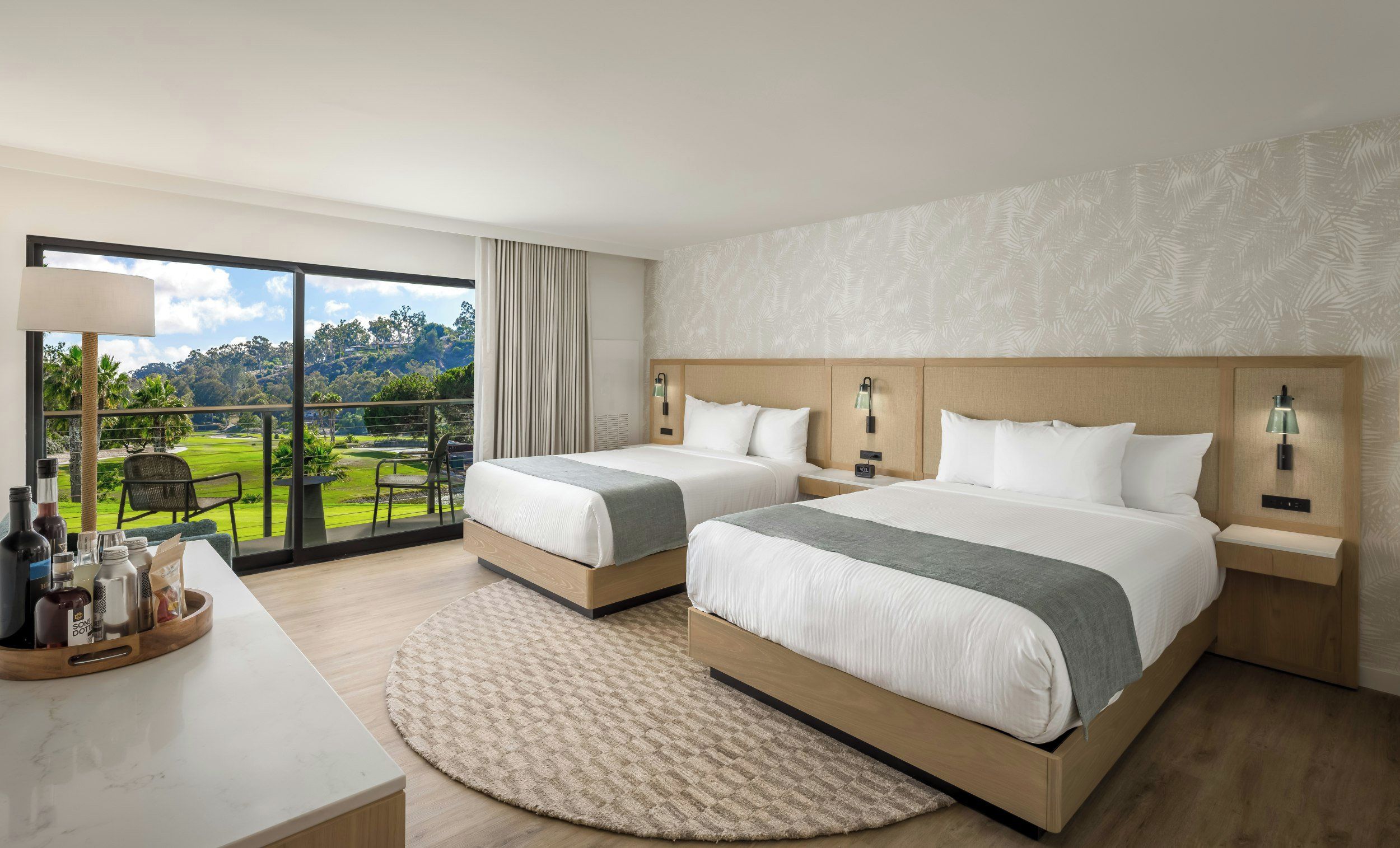 Une chambre d’hôtel avec deux lits, une grande fenêtre donnant sur un terrain de golf, un tapis rond et une petite table avec des bouteilles et des verres.