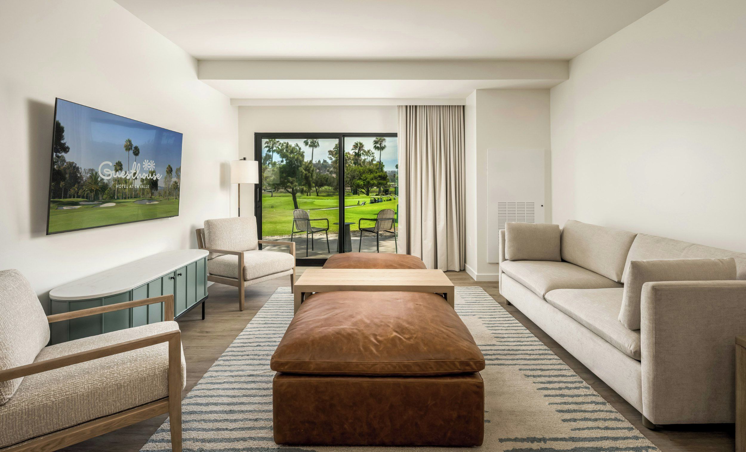 A modern hotel room with a large window offering a view of a golf course, a flat-screen TV mounted on the wall, a sofa, a chair, and a small table.
