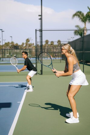 A tennis court with two players in action, a chain-link fence, and palm trees in the background. - Guesthouse Hotel at La Valle (Rancho Santa Fe)
