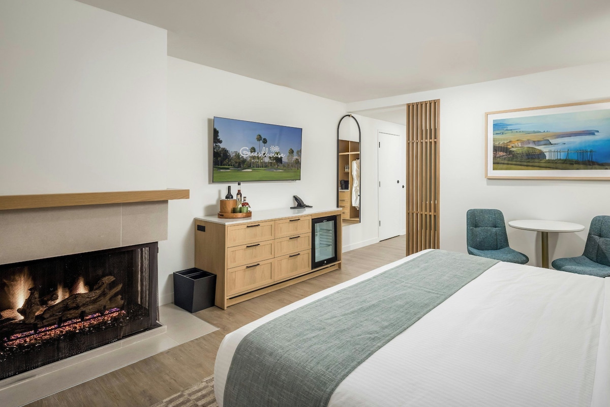 A modern hotel room with a fireplace, a bed, a desk with a computer, and two chairs.