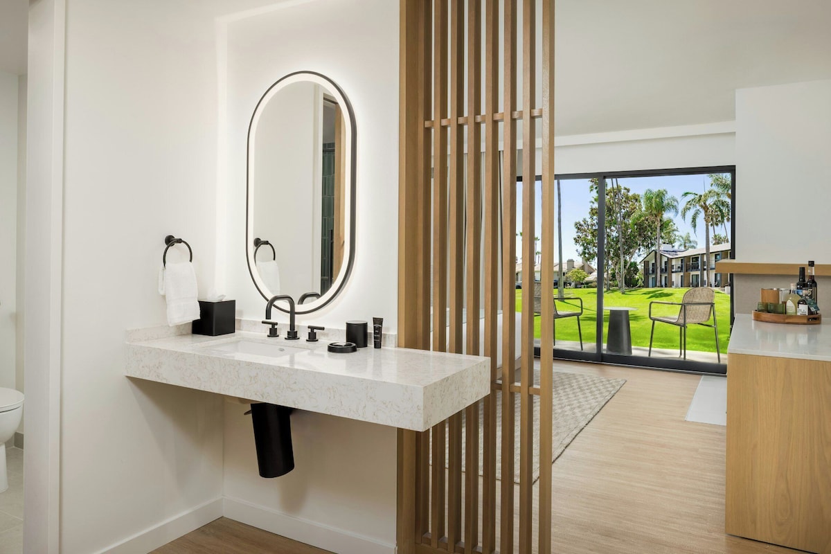 A modern bathroom with a marble sink, a large mirror, and a view of a green outdoor area through a glass door.