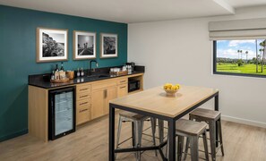 Eine moderne Küche mit einer türkisfarbenen Akzentwand, einer schwarzen Arbeitsplatte und Holzschränken. Über der Arbeitsplatte befinden sich drei gerahmte Schwarz-Weiß-Fotos. Auf dem Tisch steht eine Schüssel mit Zitronen, und ein Fenster bietet Blick auf einen Golfplatz.