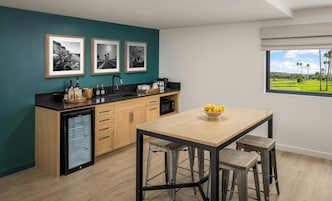 Een moderne keuken met een blauwgroene accentmuur, een zwart aanrecht en houten kastjes. Boven het aanrecht hangen drie zwart-witfoto’s in lijsten. Op de tafel staat een schaal met citroenen, en door het raam is een golfbaan te zien.