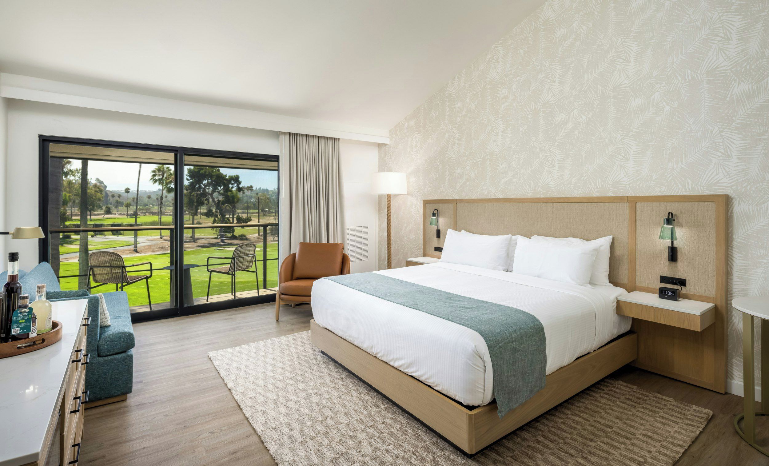 A hotel room with a large bed, a view of a golf course, a seating area with a chair, and a small table with a tray of items.
