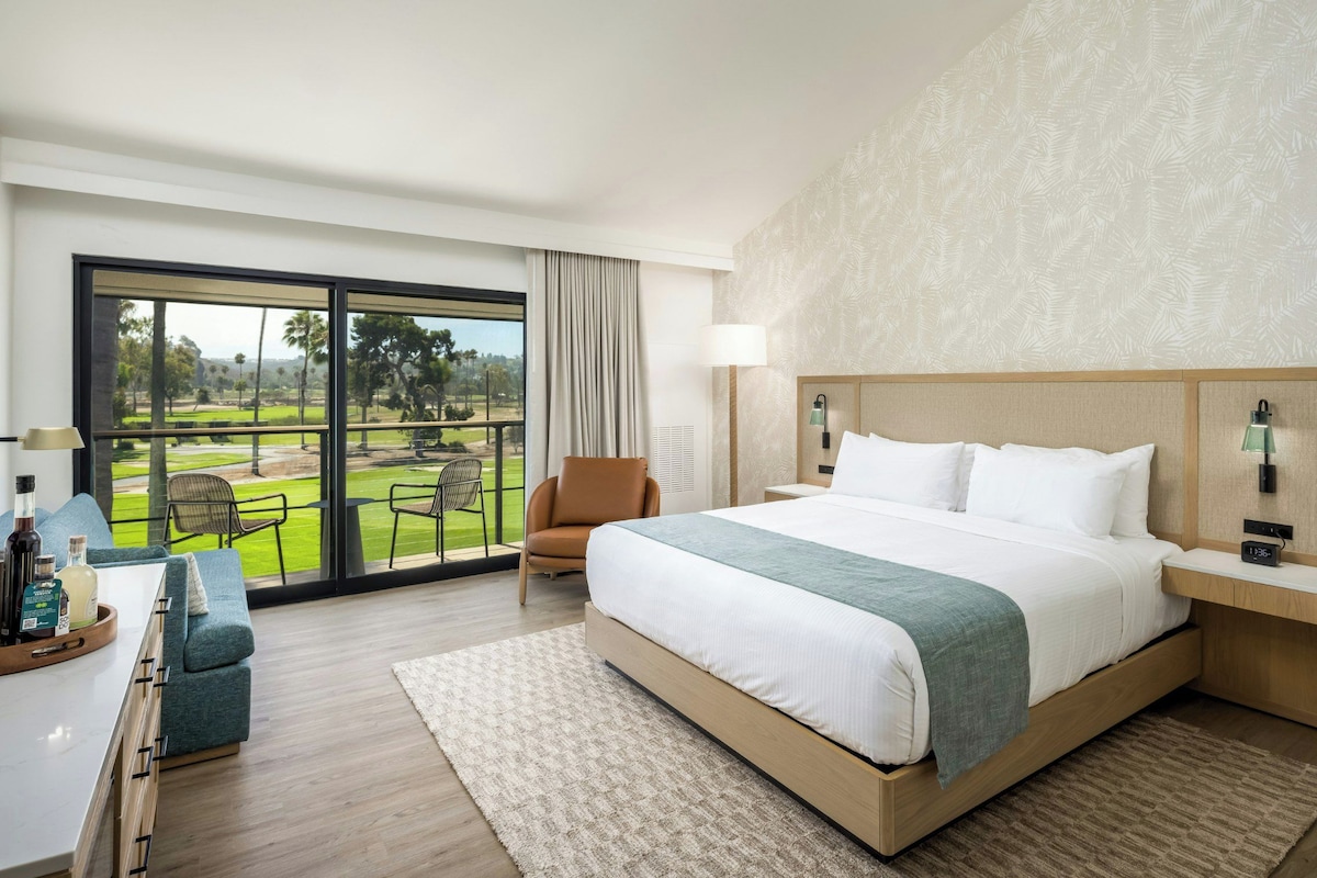A hotel room with a large bed, a view of a golf course, a seating area with a chair, and a small table with a tray of items.
