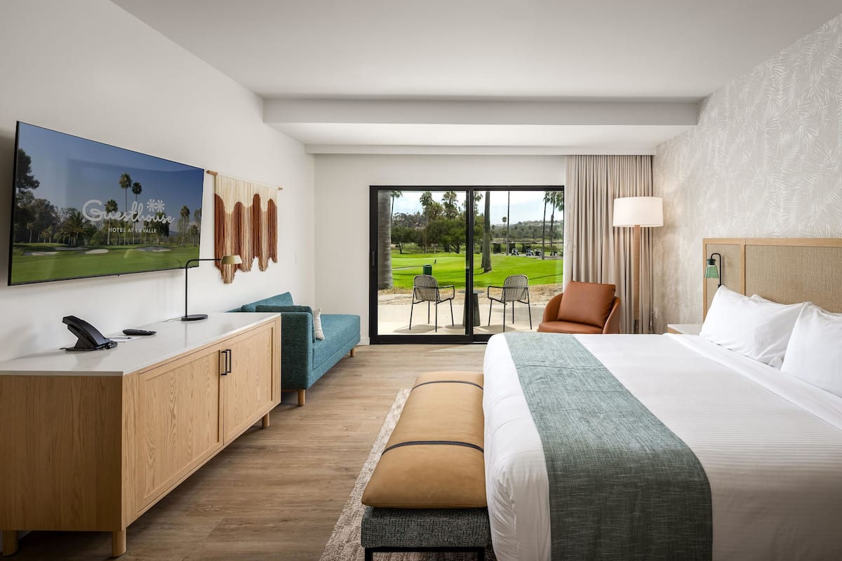 A hotel room with a large TV, a desk, a sofa, a bed, and a view of a golf course.