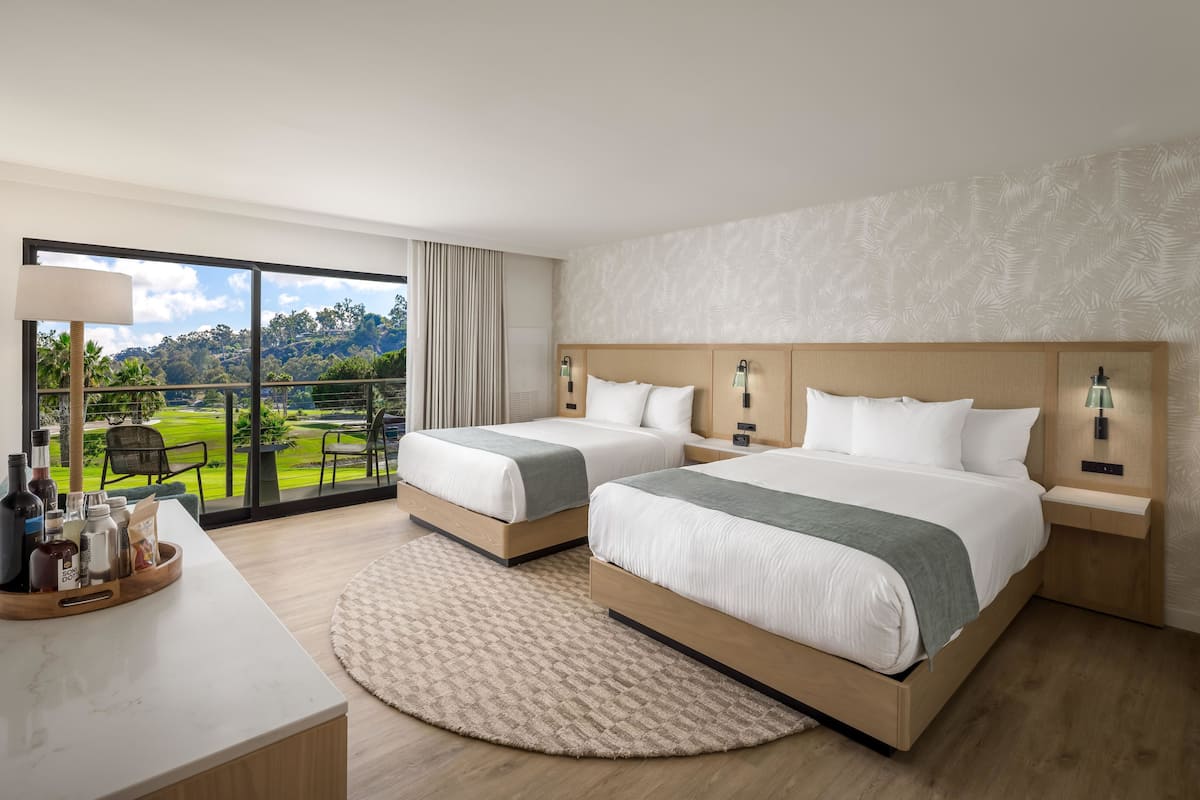 A hotel room with two beds, a large window with a view of a golf course, a desk with toiletries, and a round rug.