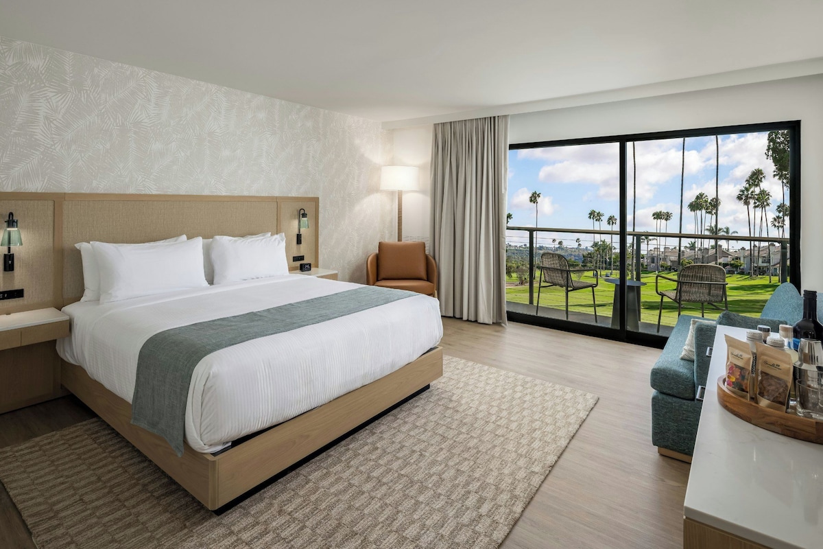 A hotel room with a large bed, a nightstand, a lamp, a chair, and a view of a golf course through the window.
